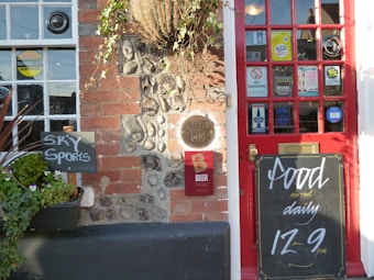 A pub entrance featuring a red door adorned with multiple stickers and signs including 'no smoking' and 'vote now'. Adjacent to the door, there is a chalkboard displaying the message 'Food served daily 12-9 pm'. Beside the entrance, a small blackboard indicates the availability of 'Sky Sports'. The wall consists of red brick and stones with a hanging plant above.