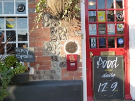 A pub entrance featuring a red door adorned with multiple stickers and signs including 'no smoking' and 'vote now'. Adjacent to the door, there is a chalkboard displaying the message 'Food served daily 12-9 pm'. Beside the entrance, a small blackboard indicates the availability of 'Sky Sports'. The wall consists of red brick and stones with a hanging plant above.