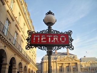 An ornate lamp post with an art nouveau style supports a red sign that says 'METRO'. The backdrop consists of classic Parisian architecture with beige stone buildings and decorative elements. The sky is clear with a soft blue hue, and a small French flag is visible on one of the buildings.