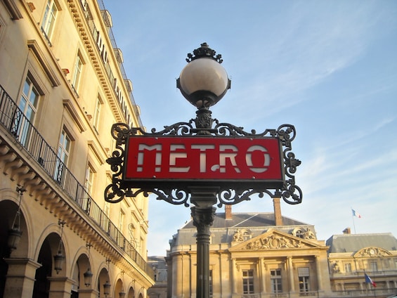 An ornate lamp post with an art nouveau style supports a red sign that says 'METRO'. The backdrop consists of classic Parisian architecture with beige stone buildings and decorative elements. The sky is clear with a soft blue hue, and a small French flag is visible on one of the buildings.