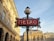 An ornate lamp post with an art nouveau style supports a red sign that says 'METRO'. The backdrop consists of classic Parisian architecture with beige stone buildings and decorative elements. The sky is clear with a soft blue hue, and a small French flag is visible on one of the buildings.