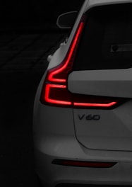A close-up view of the back of a white car, focusing on the distinct L-shaped red tail lights that are illuminated. The model designation 'V60' is visible on the car's rear. The background is dark, contrasting with the bright red lights and the white paint of the car.
