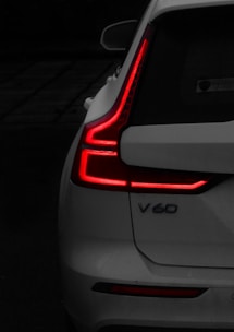 A close-up view of the back of a white car, focusing on the distinct L-shaped red tail lights that are illuminated. The model designation 'V60' is visible on the car's rear. The background is dark, contrasting with the bright red lights and the white paint of the car.