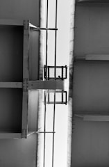 A black and white image capturing the underside of a large metallic structure, possibly a bridge, with beams and girders. There's a small platform with a mesh fence and two vertical pipes running parallel in the middle.