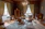 An elegantly decorated vintage living room with antique furniture. The room has two large curtained windows allowing natural light to brighten the space. A round wooden table is surrounded by upholstered chairs. A wooden wardrobe and a small table are placed against the walls, alongside a small decorative cabinet with intricate woodwork. A chandelier hangs from the wooden beamed ceiling, and a patterned rug covers part of the wooden floor.
