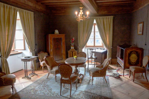 An elegantly decorated vintage living room with antique furniture. The room has two large curtained windows allowing natural light to brighten the space. A round wooden table is surrounded by upholstered chairs. A wooden wardrobe and a small table are placed against the walls, alongside a small decorative cabinet with intricate woodwork. A chandelier hangs from the wooden beamed ceiling, and a patterned rug covers part of the wooden floor.