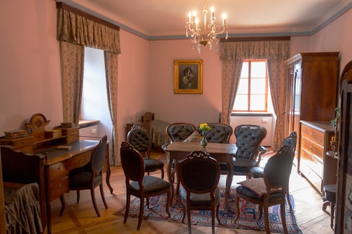 A well-lit, elegant room with classic furniture including a central wooden table surrounded by upholstered chairs with carved backs. A chandelier hangs from the ceiling, offering a warm glow. On the walls are patterned curtains framing the windows and a framed portrait painting. A desk with a curved backrest and an armoire add to the room's vintage charm. A vase with flowers sits on the table, and a detailed rug covers the floor.