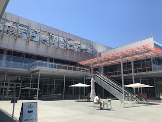 A modern outdoor plaza with a contemporary building in the background featuring large windows and a metal mesh facade. There is outdoor seating with tables and parasols, as well as a person sitting at a table. An escalator provides access to the upper level, and signage indicates a store opening.
