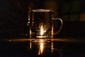 A transparent glass mug with a handle is placed on a dark surface, illuminated from within by a small, bright flame. The light creates a warm glow and casts reflections on the surface, enhancing the contrast between the light and the surrounding darkness.