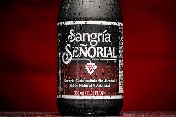 A bottle of Sangr&iacute;a Se&ntilde;orial is prominently displayed with water droplets on its surface, set against a rich red background. The label has bold white lettering, and a small image of grapes is visible in the center.