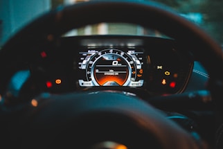 A car dashboard with an illuminated digital instrument cluster displaying speed, warning lights, and driving mode. The steering wheel partially frames the cluster, emphasizing the focus on the speedometer and indicator lights.