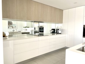 A modern kitchen features sleek white cabinets with minimalist design. There's a built-in oven, smooth countertops, and a coffee machine. The kitchen is well-lit with natural light reflecting off the glossy surfaces. A plant, framed artwork, and bowls are on display, adding a touch of decor.