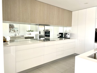 A modern kitchen features sleek white cabinets with minimalist design. There's a built-in oven, smooth countertops, and a coffee machine. The kitchen is well-lit with natural light reflecting off the glossy surfaces. A plant, framed artwork, and bowls are on display, adding a touch of decor.