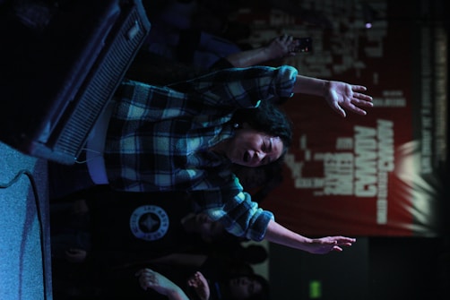 A person with their arms raised is expressing emotion in a dimly lit environment with a textured wall displaying text. The scene suggests a lively or intense atmosphere.