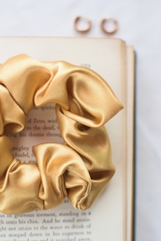 A golden satin scrunchie is placed on an open book, with two gold rings positioned above it. The page of the book has visible text but it's out of focus, emphasizing the scrunchie. The background is softly blurred, creating a gentle, serene aesthetic.