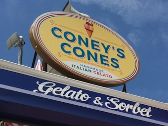 A large, oval-shaped sign for 'Coney's Cones' is prominently displayed against a clear blue sky. The sign features the text 'HANDMADE ITALIAN GELATO' with an illustration of an ice cream cone. Below the sign, there's another sign showing 'Gelato & Sorbet' in white script on a blue background.