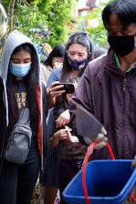 Several people are wearing masks and standing in close proximity, possibly in a queue. A younger individual is focused on a smartphone. The surroundings are lush with greenery, indicating an outdoor setting. A man in the foreground is holding a blue container with a red handle.