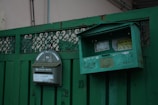 A pair of green metal mailboxes are mounted on a green fence. One mailbox has a curved top with stickers and a slot, while the other has a rectangular shape with an open lid, exposing its interior. The fence features intricate metalwork and the mailboxes have a worn, weathered appearance.