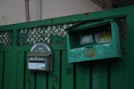 A pair of green metal mailboxes are mounted on a green fence. One mailbox has a curved top with stickers and a slot, while the other has a rectangular shape with an open lid, exposing its interior. The fence features intricate metalwork and the mailboxes have a worn, weathered appearance.