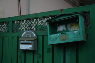 A pair of green metal mailboxes are mounted on a green fence. One mailbox has a curved top with stickers and a slot, while the other has a rectangular shape with an open lid, exposing its interior. The fence features intricate metalwork and the mailboxes have a worn, weathered appearance.