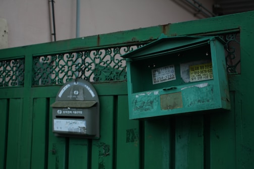 A pair of green metal mailboxes are mounted on a green fence. One mailbox has a curved top with stickers and a slot, while the other has a rectangular shape with an open lid, exposing its interior. The fence features intricate metalwork and the mailboxes have a worn, weathered appearance.