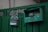 A pair of green metal mailboxes are mounted on a green fence. One mailbox has a curved top with stickers and a slot, while the other has a rectangular shape with an open lid, exposing its interior. The fence features intricate metalwork and the mailboxes have a worn, weathered appearance.