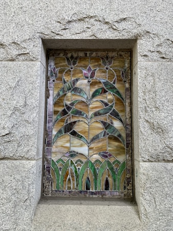 A decorative stained glass panel is embedded in a stone wall. The design features intricate patterns with floral motifs, utilizing curving lines and symmetrical shapes. The color scheme includes earthy tones like green, brown, and purple, with a textured stone frame surrounding the panel.