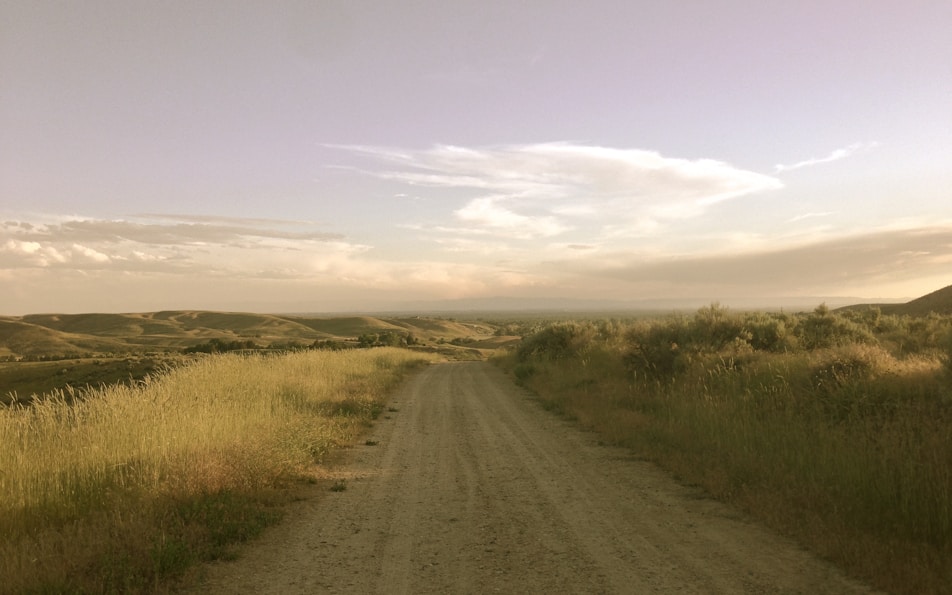 brown dirt road between green grass field under white clouds and blue sky during daytime