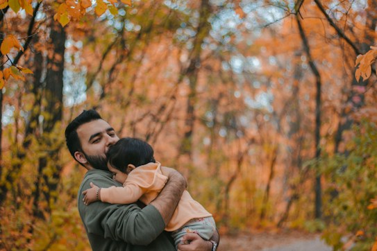 A man holding a child lovingly, surrounded by a forest with vibrant autumn colors. The leaves are bright orange and yellow, and the path is softly blurred, creating a warm and intimate atmosphere.