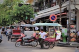A bustling street scene featuring a man sitting in a tuk-tuk parked on a roadside. Nearby, a scooter is parked, and a no-parking sign is visible above him. The background shows a busy street lined with eateries and various businesses, with trees providing shade and people walking by.