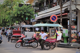 A bustling street scene featuring a man sitting in a tuk-tuk parked on a roadside. Nearby, a scooter is parked, and a no-parking sign is visible above him. The background shows a busy street lined with eateries and various businesses, with trees providing shade and people walking by.