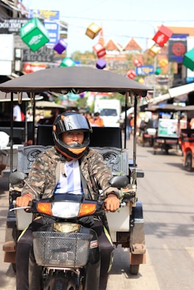 A man rides a motorcycle taxi through a busy street adorned with colorful cube decorations hanging above. The street is lined with shops and other vehicles, creating a lively urban scene. The man wears a camouflage jacket and a helmet with an orange stripe.
