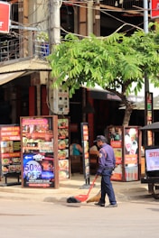 A street scene featuring a man in a uniform sweeping the ground with a broom and dustpan. Surrounding him are multiple vibrant posters and signs advertising food, beverages, and special promotions. A tree with green leaves provides some shade, and behind the man is a building that appears to house various businesses, including a restaurant or bar based on the visible menus.