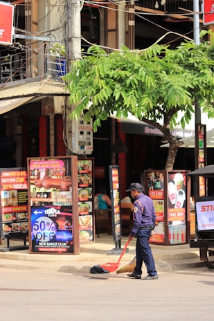 A street scene featuring a man in a uniform sweeping the ground with a broom and dustpan. Surrounding him are multiple vibrant posters and signs advertising food, beverages, and special promotions. A tree with green leaves provides some shade, and behind the man is a building that appears to house various businesses, including a restaurant or bar based on the visible menus.