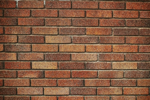 A wall constructed of evenly laid red and brown bricks, with visible mortar in between each brick. The bricks have a rough texture and vary slightly in color, adding a sense of depth and warmth to the wall.