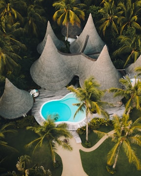 A resort-style setting with several cone-shaped thatched-roof huts surrounded by lush tropical vegetation. A vibrant blue swimming pool is centrally located on a wooden deck, with pathways winding through neatly manicured gardens.