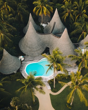 A resort-style setting with several cone-shaped thatched-roof huts surrounded by lush tropical vegetation. A vibrant blue swimming pool is centrally located on a wooden deck, with pathways winding through neatly manicured gardens.