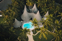 Aerial view of a tropical resort surrounded by dense palm trees, featuring uniquely designed huts with thatched, pointed roofs and a bright turquoise swimming pool at the center.
