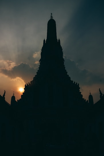 A large intricately designed temple silhouette against a backdrop of a setting or rising sun. The sky is partially covered by clouds, with rays of sunlight piercing through, creating a dramatic contrast.