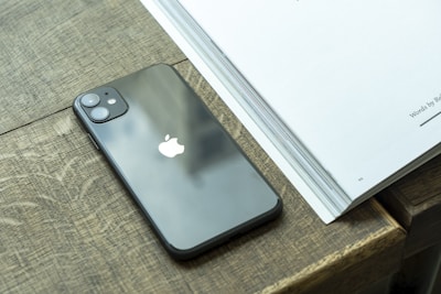 A smartphone with a sleek black finish and dual camera lenses rests on a wooden surface beside an open book with visible pages.