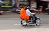 A Buddhist monk wearing vibrant orange robes rides on the back of a motorcycle driven by another person in a white shirt and jeans, who is wearing a helmet. The background is blurred, suggesting motion and a busy street environment.