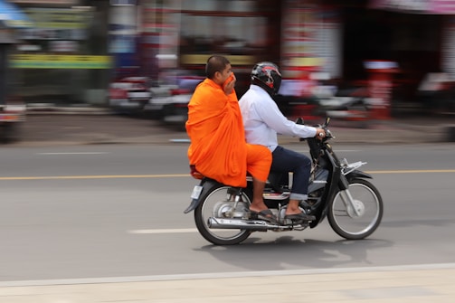 A Buddhist monk wearing vibrant orange robes rides on the back of a motorcycle driven by another person in a white shirt and jeans, who is wearing a helmet. The background is blurred, suggesting motion and a busy street environment.