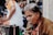 An older person with gray hair is intently speaking or singing into a microphone, with their face showing focus and emotion. They are wearing a brown jacket over an orange shirt, and the scene appears to be in a bustling outdoor market or street setting. Another individual in a white and orange shirt is sitting nearby, partially visible.