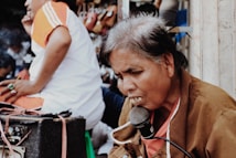 An older person with gray hair is intently speaking or singing into a microphone, with their face showing focus and emotion. They are wearing a brown jacket over an orange shirt, and the scene appears to be in a bustling outdoor market or street setting. Another individual in a white and orange shirt is sitting nearby, partially visible.