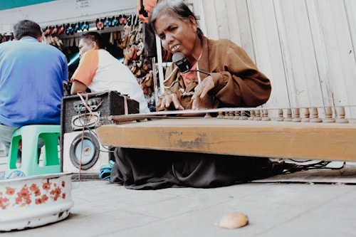 An elderly person is playing a string instrument on the street. The individual appears to be focused or singing, with a microphone in front of them. There are people seated nearby, possibly listening. Various items such as a speaker and colorful plastic stools are visible. Shoes are displayed in the background.