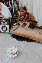 An elderly person is playing a stringed musical instrument on the street, sitting on the ground. They have a microphone attached to their neck. In the foreground, there is a bowl with some money in it, suggesting busking. A large speaker is visible next to them, and other people are seen in the background.
