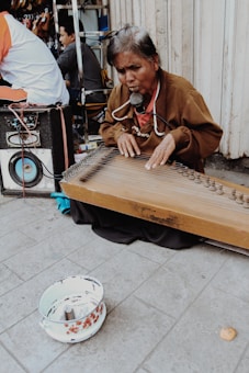 An elderly person is playing a stringed musical instrument on the street, sitting on the ground. They have a microphone attached to their neck. In the foreground, there is a bowl with some money in it, suggesting busking. A large speaker is visible next to them, and other people are seen in the background.