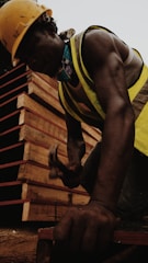A construction worker is hammering a nail into a wooden surface, wearing a yellow hard hat and reflective vest. The worker's muscular arm is visible, and in the background, there are stacked wooden boards.