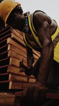 A construction worker is hammering a nail into a wooden surface, wearing a yellow hard hat and reflective vest. The worker's muscular arm is visible, and in the background, there are stacked wooden boards.