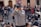 A person wearing a gray jacket and a dark cap is taking a photo with a large camera lens. They are positioned in a busy outdoor setting, surrounded by a crowd of people who are walking and gathering in groups. The background includes an old building with arched doorways.